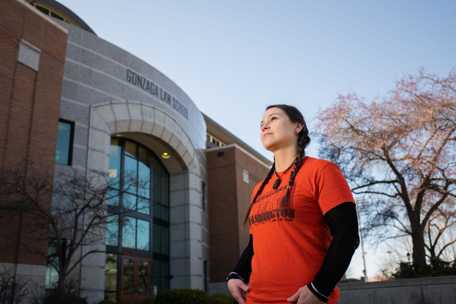 A woman with brown hair in two braids looking off into the distance. She's wearing an orange shirt and standing in front of the Gonzaga Law School building.