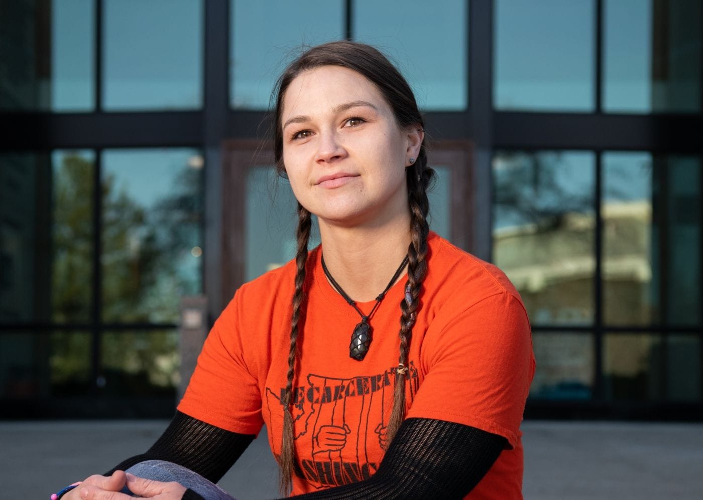A woman with brown hair in two braids smiling slightly to the camera. She's wearing an orange shirt that says "Decarcerate Washington."
