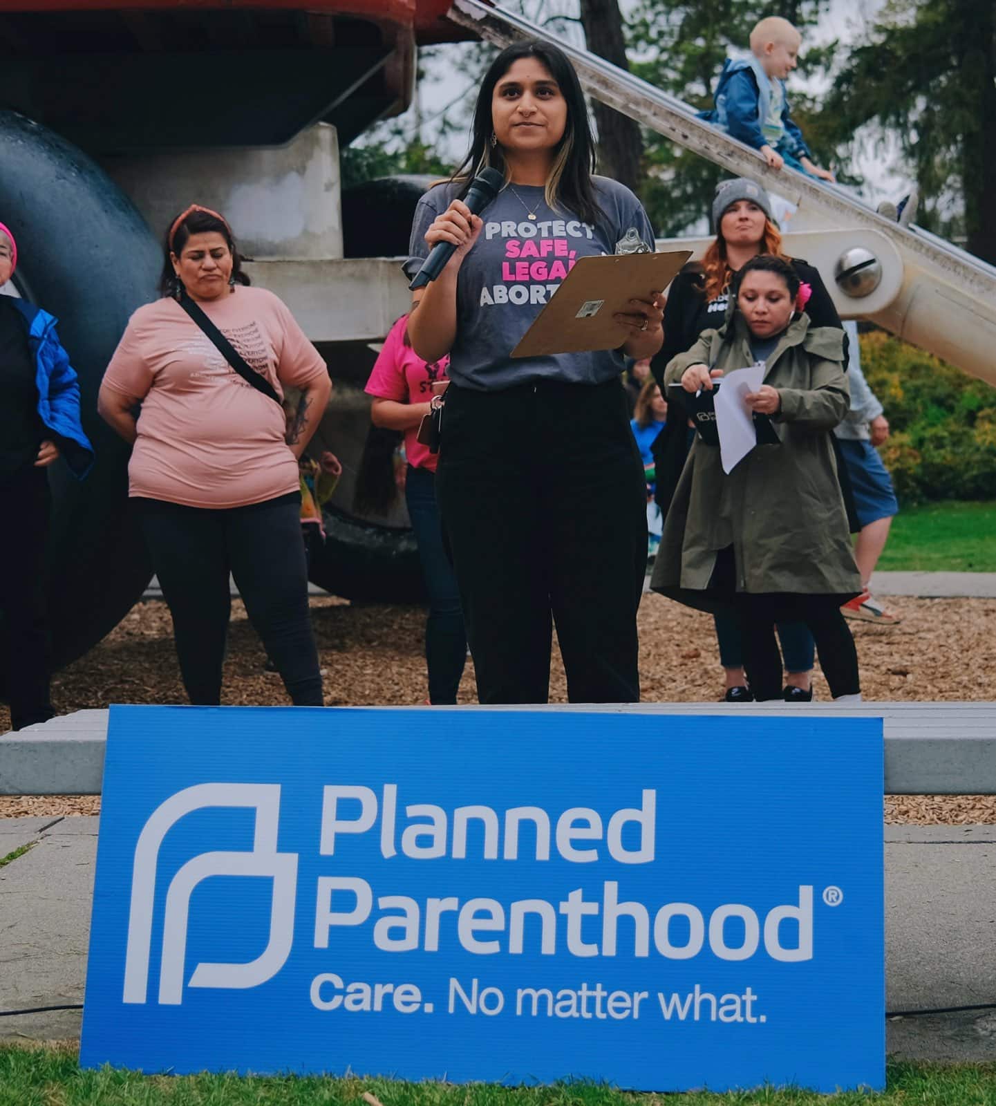 Sarah Dixit is speaking at a Planned Parenthood rally behind a sign that reads, “Planned Parenthood. Care. No matter what.” Dixit wears a shirt that reads “Protect Safe, Legal abortion.”